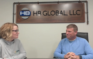 A photo of Stacy (left) and Bob (right) sitting at a table in an HB Global conference room talking to each other.