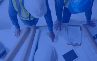 A photo of three people gathered around blueprints and schematics in yellow vests and hard hats collaborating together.